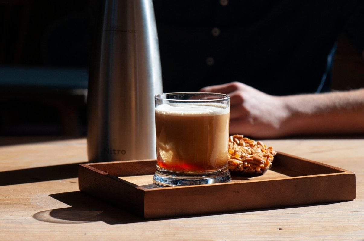 Gals mit schaumigem Nitro Coffee und Nusskeks auf Holztablet, dahinter ein Teil von einem Sahnespender und ein Teil von einem Arm