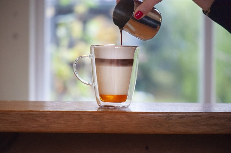 in ein Glas mit Honig am Boden und Milchschaum wird von oben ein Espresso eingegossen