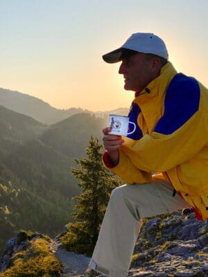 Kaffeepause nach dem Wandern bei Sonnenaufgang – Mann mit Emaille-Tasse auf Berggipfel, Symbol für Kaffee & Outdoor-Genuss im Sportkontext