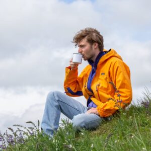 Mann in orangefarbener Outdoorjacke sitzt in einer Blumenwiese und genießt Kaffee aus einer Emailletasse – Naturmoment mit Specialty Coffee.