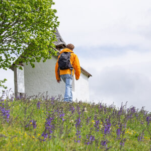 Mann wandert auf einen Berggipfel mit kleiner Kapelle in orangener Jacke und Kaffee im Rucksack